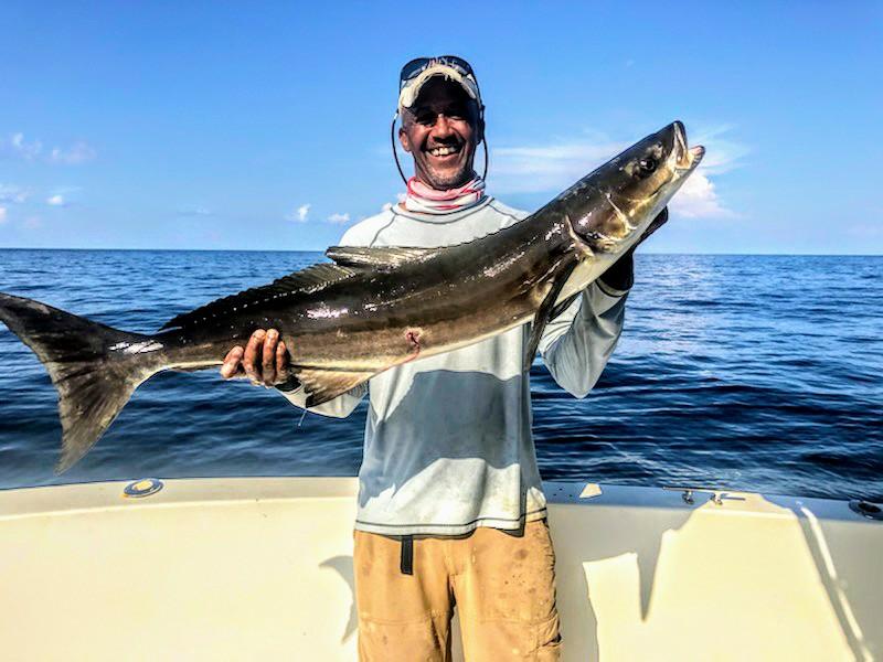 Smiling angler holding a large Cobia fish on a boat deck with blue ocean background