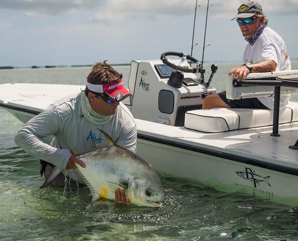 Experienced fishing guide holding a large permit fish in shallow water next to a flats boat with captain at helm in Key West