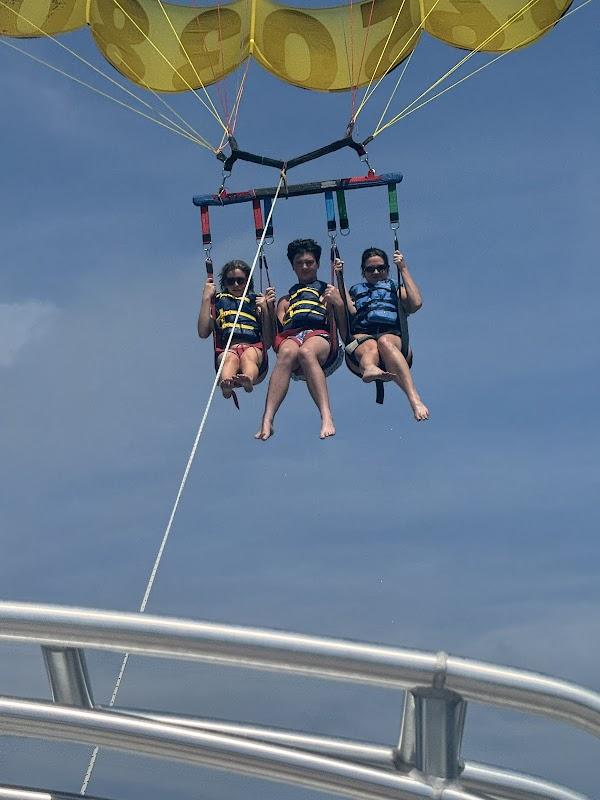 Three guests enjoying a tandem parasailing ride with a yellow smiley-face parachute under a clear blue sky
