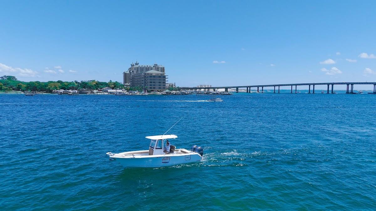 1st Choice Charters center console fishing boat cruising through Destin Harbor with the Destin Bridge and Emerald Grande resort in the background under a blue sky.