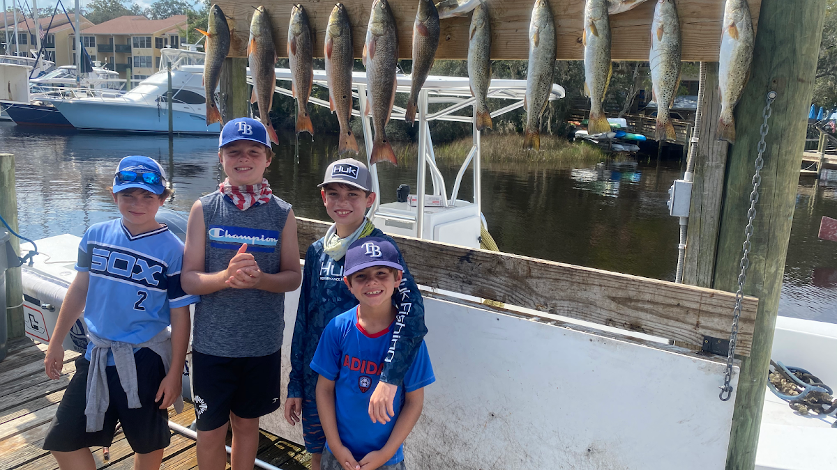 Four smiling boys standing on a dock with a large catch of redfish and trout hanging on a rack behind them at Last Cast Charters in Destin, FL.